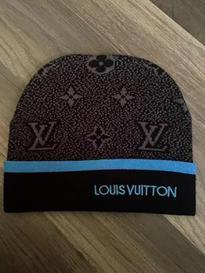 Svart mössa med blå kant - Snygg svart mössa  med klassiskt monogrammönster i grått och en blå kant. Märkets logga är broderad i blått framtill. Perfekt accessoar för dig som vill ha en exklusiv och trendig look.