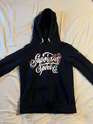 Mörkblå Superdry hoodie med blommor - Snygg mörkblå hoodie från Superdry med stor huva i storlek S. Framsidan har ett färgglatt tryck med texten 'Superdry Spirit' dekorerat med blommor i rosa, gult och blått. Nypris: 699kr / Mitt pris: 399kr , Hör av er vid funderingar 🤝⭐️