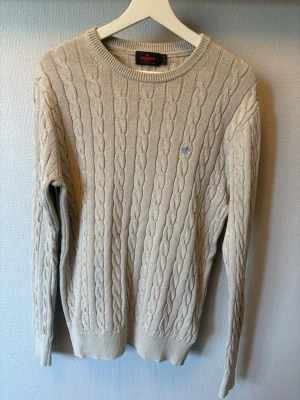 Beige kabelstickad tröja Morris - Kabelstickad beige tröja från Morris med rund halsringning och diskret blå brodyr på bröstet. Tröjan har långa ärmar och ribbade muddar vid ärmslut och nederkant. Perfekt för lager-på-lager och klassisk stil. Skick 9/10 använd endrast fåtal gånger