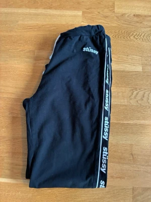 Svarta mjukisbyxor från Stüssy - Säljer ett par svarta mjukisbyxor från Stüssy med vita loggor längs sidorna och broderad logga vid fickan. Byxorna har en avslappnad passform och resår i midjan. Perfekta för chill dagar eller streetwear-stil.