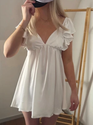 Vit kort klänning med volangärmar - Söt vit kortklänning med djup v-ringning och volangärmar. Klänningen har en luftig passform och är tillverkad i ett mjukt, lätt tyg som faller fint. Perfekt för sommar eller studenten. 