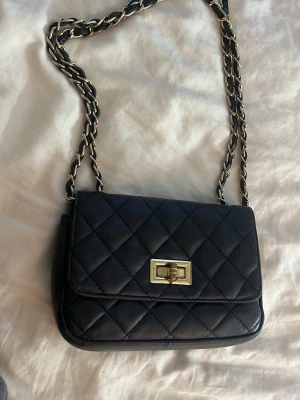 Svart quiltad axelväska med kedjerem (båda axelväska och handväska) - Stilren svart axelväska i quiltad läderimitation med guldfärgad vridknäppe framtill. Kedjerem med insytt läder ger en exklusiv vibe. Köpte på en kryssningsbåt för 599. Lite använd men lika snygg och funkar exakt som när den köptes. 