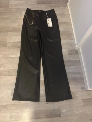 Svarta vida byxor i skinnimitation - Snygga svarta byxor i skinnimitation från ONLY med vida ben och hög midja i stk M. Byxorna har guldfärgade detaljer och en kedja framtill som extra detalj. Perfekta för dig som vill sticka ut med en trendig look. Aldrig användna. 