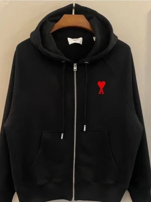 Svart hoodie med röd Ami-de-Coeur - Svart hoodie från AMI Paris med dragkedja, stor huva och känguruficka. På bröstet sitter det ikoniska röda Ami-de-Coeur-broderiet. Tillverkad i mjuk bomull för en skön och avslappnad look. Perfekt för dig som gillar stilrena detaljer. 