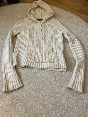 Beige stickad hoodie Brandy Melville - Stickad hoodie i beige från Brandy Melville med kabelstickat mönster, knappdetaljer vid halsen och fickor framtill. Tröjan har långa ärmar och ribbade muddar. Perfekt för mysiga dagar och enkel att styla till jeans eller kjol.