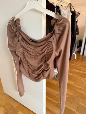 Brun långärmad topp från Shein - Trendig brun långärmad topp från Shein med rynkade detaljer framtill och volang vid axlarna. Toppen har en croppad passform och är tillverkad i ett mjukt, stretchigt material. Perfekt för att lyfta din outfit med en cool touch.