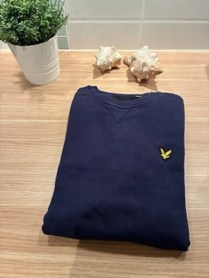 Mörkblå sweatshirt från Lyle & Scott - Mörkblå sweatshirt från Lyle & Scott med klassisk rund halsringning och gul logga broderad på bröstet. Tröjan har långa ärmar och ribbade muddar vid ärmslut och nederkant. Perfekt för en stilren och avslappnad look.