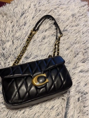 Svart quiltad handväska från Coach - Snygg svart quiltad handväska från Coach med guldigt kedjeaxelband och stort C-spänne i metall på framsidan. Väskan är tillverkad i läder och har en klassisk rektangulär form med lock. Perfekt för dig som gillar stilrena och lyxiga accessoarer.