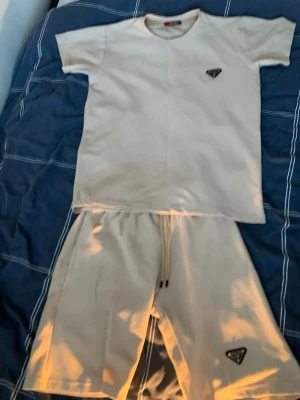 Beige set från Prada med logga - Säljer ett beige set från Prada med matchande t-shirt och shorts. Båda plaggen har den ikoniska svarta Prada-loggan framtill. Materialet är mjukt och har en lätt ribbad struktur. Perfekt för dig som gillar exklusiva och stilrena underkläder. Riktigt bra för sommaren som är påväg.