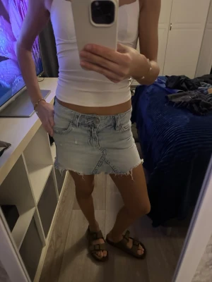 Ljusblå jeanskjol med fransig kant - Trendig ljusblå jeanskjol med rå fransig kant och klassiska fickor fram och bak. Kjolen har en kort passform och stängs med dragkedja och knapp framtill.