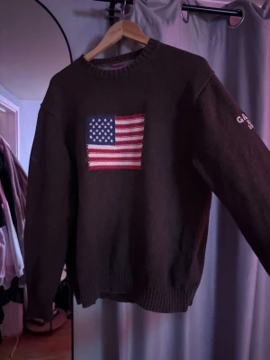 Mörkbrun stickad GANT-tröja USA-flagga - Stickad mörkbrun tröja från GANT med amerikansk flagga broderad på bröstet och vit GANT Est. 1949-text på ärmen. Rund halsringning och ribbade muddar vid ärmslut och nederkant. Perfekt för dig som gillar klassisk preppy-stil.