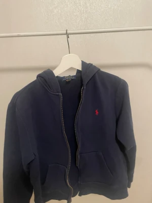 Mörkblå hoodie från Polo Ralph Lauren - Mörkblå hoodie från Polo Ralph Lauren med dragkedja framtill och klassisk röd logga broderad på bröstet. Tröjan har huva och två fickor framtill. Tillverkad i mjukt bomullsmaterial som är skönt att bära. Perfekt för en avslappnad och stilren look.