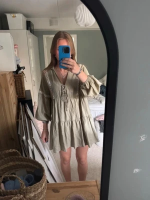 Beige kortklänning med snörning - Säljer en beige kortklänning med volang och snörning framtill. Klänningen har v-ringning, vida långärmar och spetsdetaljer längs axlar och ärmar. Perfekt för dig som gillar en avslappnad och bohemisk stil.