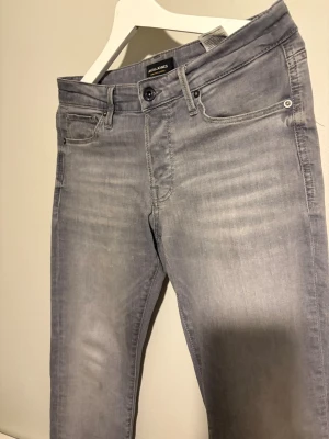 Grå slim fit jeans från Jack & Jones - Snygga grå slim/glenn jeans från Jack & Jones med klassisk femficksdesign och diskreta slitningar framtill. Jeansen har normal midja och raka ben, tillverkade i mjuk denim med lite stretch för extra komfort. Knappt använda W29 L32. Skriv vid funderingar 