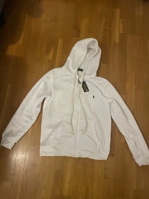 Vit hoodie från Polo Ralph Lauren - Säljer en fräsch vit hoodie från Polo Ralph Lauren med dragkedja, huva med snören och två fickor framtill. Klassisk liten logga broderad på bröstet. Perfekt för dig som gillar stilrena och bekväma plagg.