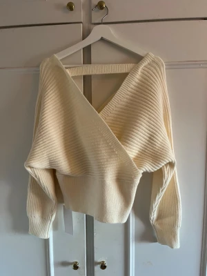 Krämvit omlottstickad tröja - Säljer en krämvit stickad tröja med omlottdesign och bred ribbad mudd i midjan. Tröjan har långa ärmar och är tillverkad i ett mjukt, ribbstickat material. Perfekt för dig som gillar en avslappnad men ändå trendig look. 