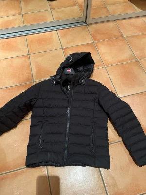 Svart pufferjacka från colmar - Säljer en svart pufferjacka från colmar med huva och dragkedja framtill. Jackan har två sidofickor med dragkedja och är fylld med naturligt dun för extra värme. Snygg, quiltad design som passar perfekt till kalla vinterdagar.