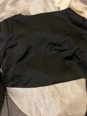 Svart croppad bolerotopp  - Snygg svart croppad bolerotoppi ett glansigt, mjukt material. Toppen har långa ärmar och en enkel, stilren design som passar perfekt till många olika outfits. Materialet känns som satin eller polyester och ger en lyxig känsla.