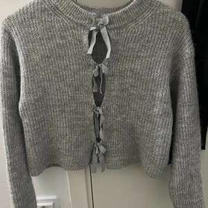 Grå stickad kofta med knytband - Supermysig grå stickad kofta med croppad passform och långa ärmar. Framsidan har flera snygga knytband som ger en unik look. Perfekt att styla med jeans eller kjol för en chill vibe. Ribbad kant vid halsen och mjukt material som känns skönt mot huden.