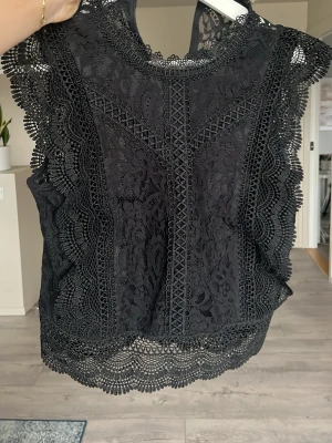 Svart spetsblus med volangdetaljer - Supersnygg svart blus i spets med vackra volangdetaljer längs ärmar och nederkant. Blusen är ärmlös och har ett feminint mönster i spetsen som ger en elegant look. Perfekt att styla med smycken för en trendig outfit.