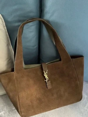 Brun mocka handväska från YSL - Säljer en snygg brun handväska i mocka från Yves Saint Laurent. Väskan har en stilren, fyrkantig form och pryds av det klassiska YSL-emblemet i guld framtill. Rymlig insida och breda axelband gör den både trendig och praktisk. 👜💌✨