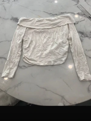 Vit offshoulder långärmad topp - Trendig vit offshoulder topp med långa ärmar och bred krage. Toppen har en lätt croppad passform och är tillverkad i mjukt bomullsmaterial. Perfekt för dig som vill ha en stilren och enkel look.