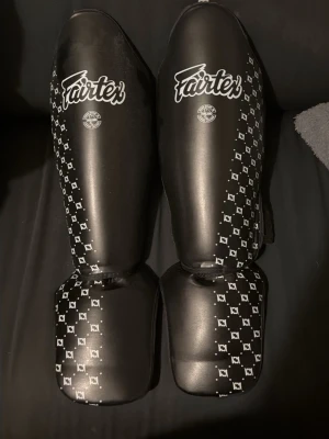 Svarta benskydd Fairtex - Säljer ett par svarta benskydd från Fairtex med vit logga och mönster längs sidorna. Benskydden har justerbara kardborreband för bra passform och är tillverkade i slitstarkt syntetmaterial, perfekta för kampsportsträning. Benskydden är i ny skick och visar ingen användning. Ny pris 1.200kr och för att tillägga är fairtex klassat som en av dom bästa kampsport märkerna. Storlek M