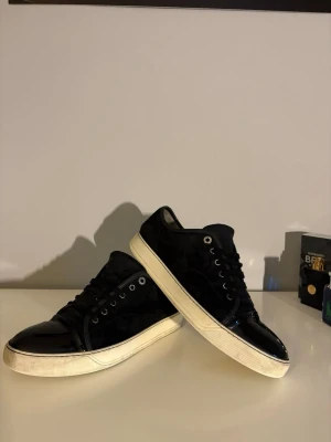 Svarta lanvins  - Snygga svarta sneakers med vit sula och svarta skosnören. Ovandelen är i en mix av blankt och matt material, vilket ger en cool kontrast. Metallfärgade öljetter och klassisk låg modell gör dem perfekta till streetwear-stilen.