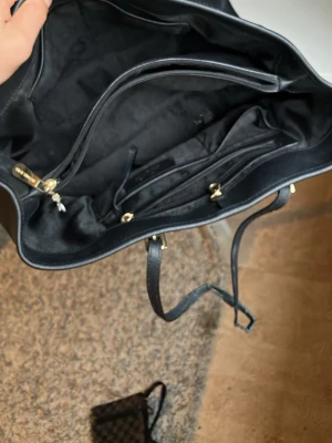 Svart shoppingväska med gulddetaljer - Stilren svart shoppingväska i konstläder med dubbla handtag och guldfärgade metalldetaljer. Väskan har rymligt huvudfack, innerficka med dragkedja och flera mindre fack för bra organisering. Perfekt för dig som vill ha plats för allt i vardagen.