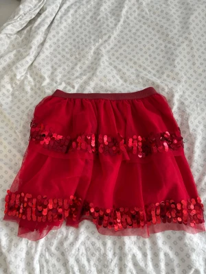 Röd kortkjol med paljetter - Snygg röd kortkjol med två rader stora paljetter och volangdetaljer. Kjolen har ett elastiskt midjeband och är gjord i ett tunt, lätt material som ger en fluffig look. Stretchigt material och stor i storleken, passar mig som är 14 år. Perfekt för fest, maskerad eller glitterfest✨💖