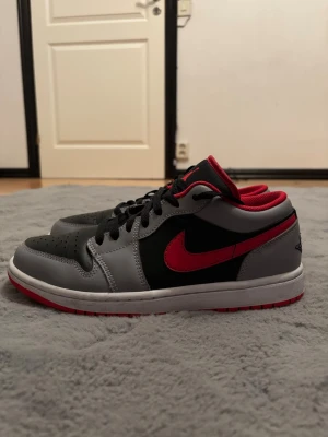 Nike Air Jordan 1 Low svart/röd/grå - Snygga Nike Air Jordan 1 Low sneakers i svart, grått och rött. Skorna har klassisk låg siluett, svart läder på tå och sidor, grått läder runtom och röd swoosh samt detaljer. Air Jordan-logga på hälen och röd yttersula. Perfekta för dig som gillar streetwear.