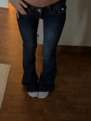 Mörkblå bootcut jeans True Religion - Säljer ett par mörkblå bootcut jeans från True Religion med snygga kontrastsömmar och detaljerade bakfickor med vita knappar. Klassisk femficksmodell i stretchigt denimtyg som sitter snyggt över höfterna och har utsvängda ben.