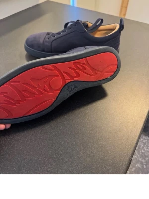 Svarta Louboutin sneakers med röd sula - Snygga svarta sneakers från Christian Louboutin i mocka med den ikoniska röda sulan. Lågt skaft och klassisk snörning. Insidan är beige och skon har en stilren, minimalistisk design som sticker ut med den röda undersidan. OBS har jättelitet hål och är kopia