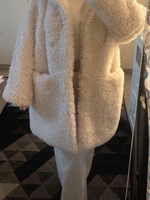 Beige oversized teddyjacka - Mysig och oversized teddyjacka i beige med stora fickor framtill och bred krage. Jackan har en fluffig och mjuk struktur som ger en trendig look. Perfekt för kalla dagar när du vill vara både varm och snygg.