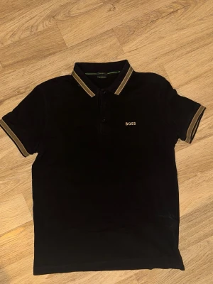 Svart piké från BOSS med gulddetaljer - Snygg svart pikétröja från BOSS med guldfärgade detaljer på krage och ärmslut. Klassisk pikékrage med knappar framtill och BOSS-logga broderad på bröstet. Tillverkad i bomull för en skön känsla och stilren look.