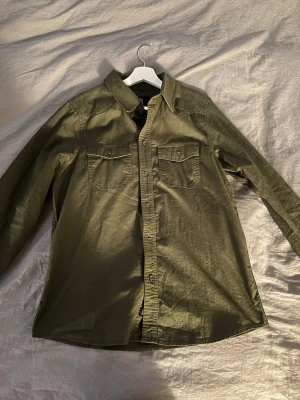 Olivgrön overshirt - Snygg olivgrön skjorta i bomull med två bröstfickor och knappar framtill. Klassisk krage och långärmad modell som passar perfekt till jeans eller chinos. Enkel och stilren design för en avslappnad look.
