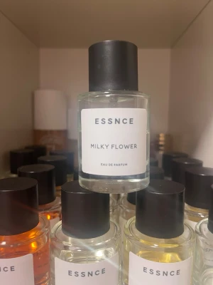 ESSNCE Milky Flower Eau de Parfum - Fräsch parfym från ESSNCE med doften Milky Flower. Flaskan är rund och genomskinlig med svart lock och vit etikett. Modern och stilren design som passar perfekt på parfymhyllan. Innehållet är klart och flaskan är gjord av glas.