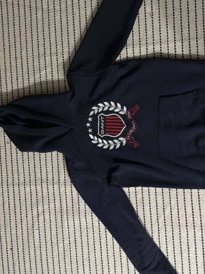 Mörkblå Gant hoodie med logga - Mörkblå hoodie från Gant med stort tryck på bröstet i vitt, rött och blått. Klassisk huva och känguruficka framtill. Tröjan har långa ärmar och är gjord i ett mjukt material som känns bekvämt. Perfekt för en sportig och avslappnad look.