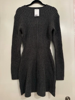 Mörkgrå ribbad stickad klänning - Mysig mörkgrå stickad klänning med ribbat mönster och lång ärm. Klänningen har en figurnära passform upptill och lätt utsvängd kjol. Perfekt för kyligare dagar och enkel att styla med boots eller sneakers.