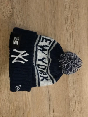 New Era New York Yankees mössa - Mörkblå och vit ribbstickad mössa från New Era med New York Yankees-logga framtill och texten 'NEW YORK YANKEES' runt om. Mössan har en stor boll i toppen i matchande färger och bred uppvikt kant. Perfekt för dig som vill ha sportig streetstyle.