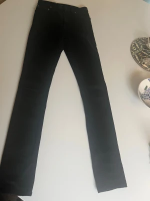 Svarta skinny jeans från Nudie Jeans - Säljer ett par svarta skinny jeans från Nudie Jeans. De har klassisk femficksdesign, dragkedjegylf och är tillverkade i stretchigt denimtyg för en tajt passform. Perfekta för dig som gillar en stilren och enkel look.
