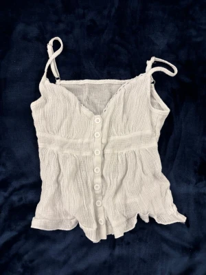 Vit topp med knappar - Söt vit linnetopp med knappar framtill och smala axelband. Toppen har en lätt rynkad struktur och volangkant nertill, vilket ger en luftig och somrig känsla. Perfekt för varma dagar och enkel att matcha med jeans eller kjol.