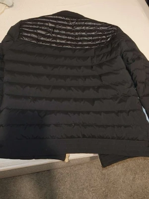 Svart dunjacka från Moncler - Snygg svart dunjacka från Moncler med quiltad design och diskret Moncler-logga på ärmen. Jackan har en klassisk passform och är tillverkad i ett glansigt material upptill och matt tyg nedtill. Perfekt för kalla vinterdagar.