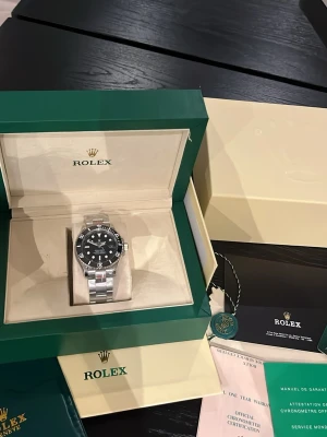 Rolex Submariner svart urtavla - Klassisk Rolex Submariner med svart urtavla och silverfärgad länk i rostfritt stål. Klockan har en rund boett, tydliga index och vridbar bezel. Levereras i originalbox med certifikat och tillbehör. Perfekt för dig som gillar exklusiva accessoarer.