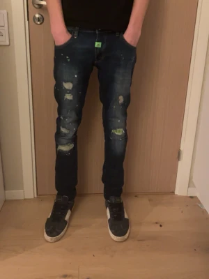 Mörkblå Philip plein jeans med färgstänk - Säljer ett par mörkblå jeans med skinny fit och slitningar på både fram- och baksida. Jeansen har coola färgstänk och patchdetaljer, samt klassiska fem fickor. Perfekta för dig som gillar en streetwear-look.