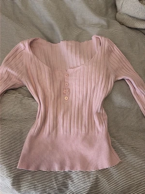ELENA GILBERT Ljusrosa ribbad stickad topp - Säljer en ljusrosa stickad topp med ribbad struktur och v-ringning. Toppen har tre knappar framtill och långa ärmar. Perfekt för dig som gillar en feminin och trendig stil.