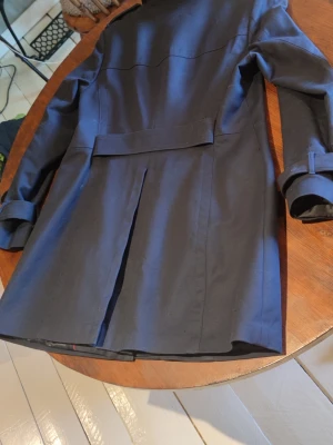 Mörkblå trenchcoat med bälte - Stilren mörkblå trenchcoat från Vero Moda med dubbelknäppning, bälte i midjan och spännen vid ärmsluten. Klassisk krage och slits bak för extra rörelsefrihet. Perfekt för dig som vill ha en tidlös och snygg jacka till vardagslooken.