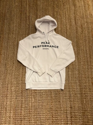 Vit hoodie Peak Performance - Snygg vit hoodie från Peak Performance med svart text på bröstet. Klassisk modell med huva samt känguruficka framtill. Tillverkad i mjukt material som känns skönt mot huden. Perfekt för en avslappnad och sportig stil.