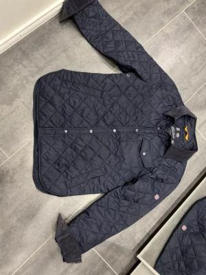 Mörkblå quiltad jacka från Morris - Quiltad mörkblå jacka från Morris med klassisk krage i manchester, bröstficka med knapp och snyggt rutmönster. Jackan har tryckknappar framtill och en patch med brittisk flagga på ärmen. Perfekt för dig som gillar stilren och tidlös look.Använd fåtals gånger.