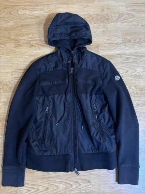 Svart Moncler cardigan tricot - Pris kan diskuteras Snygg svart vindjacka från Moncler med huva och dragkedja framtill. Jackan har två framfickor med dragkedja och Moncler-logga på ärmen. Materialmix med polyester och bomull ger en sportig look. Perfekt för dig som vill ha en stilren jacka med streetkänsla.